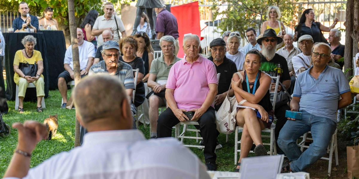 Açık havada, yeşil bir bahçede düzenlenen bir etkinliğe katılan ve sandalyelerde oturan çeşitli yaş gruplarından oluşan bir izleyici grubu. Ön planda, arkası dönük bir konuşmacı dinleyicilere hitap ederken, izleyiciler arasında pembe polo yaka tişörtlü bir adam, yanında siyah askılı bluz giyen bir kadın ve mavi tişörtlü gözlüklü bir adam dikkat çekiyor. İzleyicilerin bir kısmı dikkatle dinlerken, sol tarafta çimlerin üzerinde yürüyen bir kedi ve arka planda ayakta duran diğer katılımcılar görülüyor.
