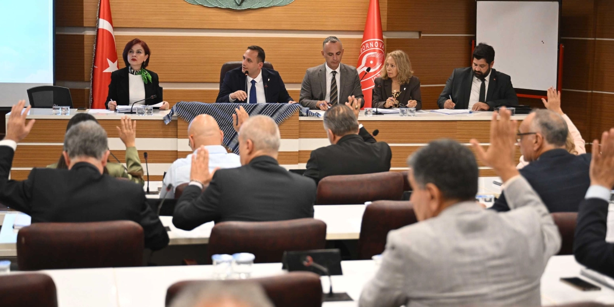 Bornova Belediye Meclisi toplantısında, meclis üyeleri oylama yapmak üzere ellerini kaldırmış durumda. Salonun ön kısmındaki protokol masasında oturan yetkililerin önünde, Filistin'i simgeleyen siyah beyaz desenli bir kefiye serili halde duruyor. Arka planda Türk bayrağı ve Bornova Belediyesi'nin logosunun bulunduğu bayrak yer alıyor.