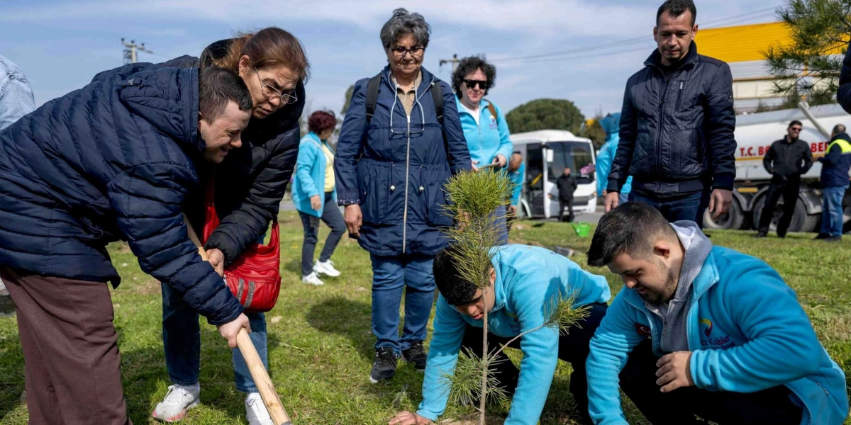 Açık havada, çimenlik bir alanda bir grup insan fidan dikiyor. Ön planda, üzerinde 