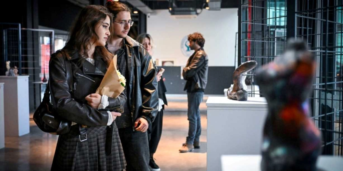 Modern bir sanat galerisinde, siyah deri ceket giymiş genç bir kadın ve yanında duran gözlüklü bir erkek, sergilenen heykelleri inceliyor. Kadın, elinde kraft kağıdına sarılı bir buket çiçek tutuyor. Arka planda ise başka ziyaretçiler galeriyi gezmeye devam ediyor.