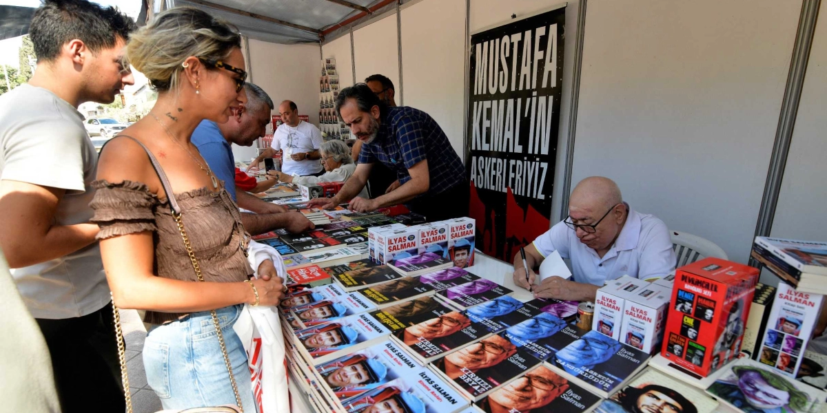 Bir kitap fuarı standında, oyuncu İlyas Salman okurları için kitaplarını imzalıyor. Masanın üzerinde İlyas Salman’ın 
