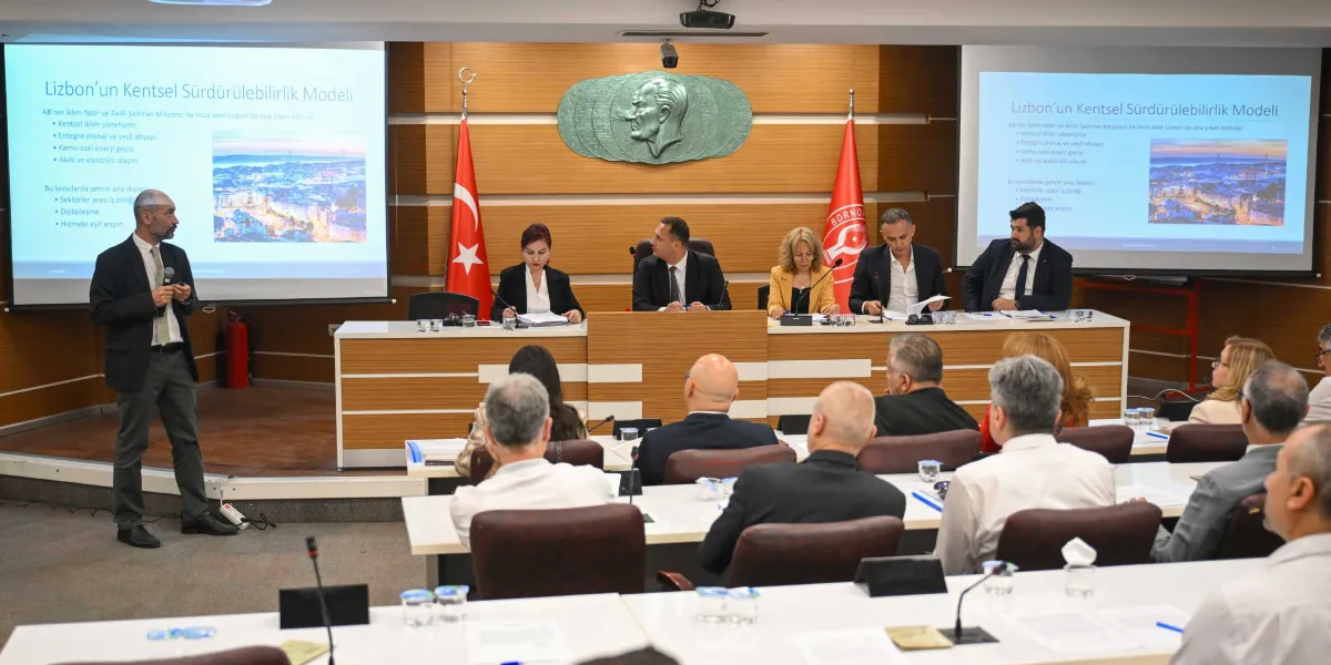 Bornova Belediyesi'nde düzenlenen bir toplantıda, kürsüde sunum yapan bir konuşmacı ve onu dinleyen katılımcılar yer alıyor. Arka planda iki ayrı projeksiyon ekranında 