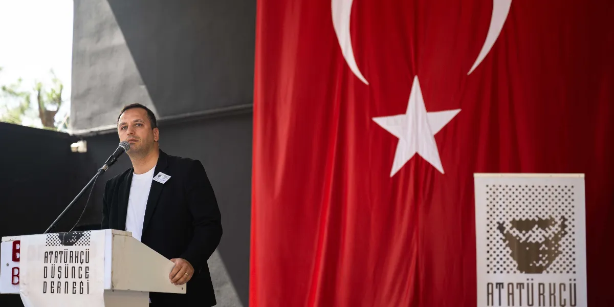 Atatürkçü Düşünce Derneği kürsüsünde konuşma yapan bir erkek, arka planda asılı büyük Türk bayrağı ve üzerinde derneğin logosu bulunan kürsü önü panelleriyle birlikte görülüyor.