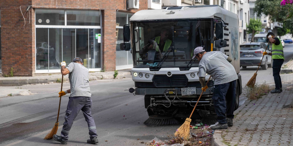 Bornova Belediyesi logolu gri tişörtler ve iş kıyafetleri giyen üç temizlik işçisi, cadde üzerinde süpürgelerle temizlik yapıyor. İşçilerden ikisi yol kenarındaki çöpleri süpürürken, üçüncüsü kaldırımda çalışıyor. Ortada, üzerinde Bornova Belediyesi yazısı bulunan büyük bir yol süpürme aracı yer alıyor.