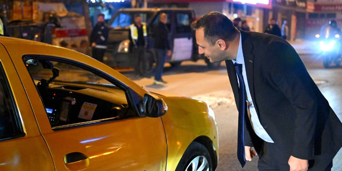 Gece vakti bir sokakta, takım elbiseli ve kravatlı bir adam, durdurduğu sarı ticari taksinin şoförüyle konuşmak için camdan içeri doğru eğilmiş bakıyor. Arka planda polis yeleği giymiş görevliler ve bir polis aracı bulunuyor.