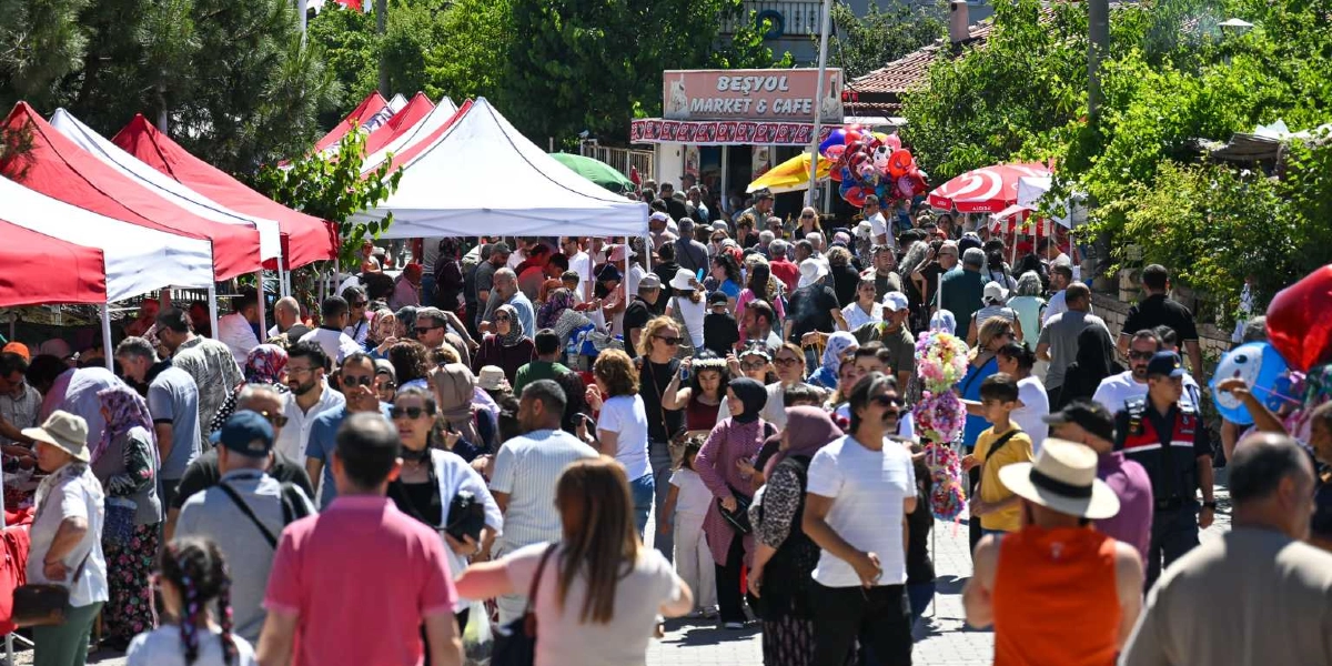 Bornova'da düzenlenen kiraz festivalinde, yan yana dizilmiş kırmızı beyaz çadırların altında alışveriş yapan ve yürüyen kalabalık bir insan grubu görülüyor. Arka planda 