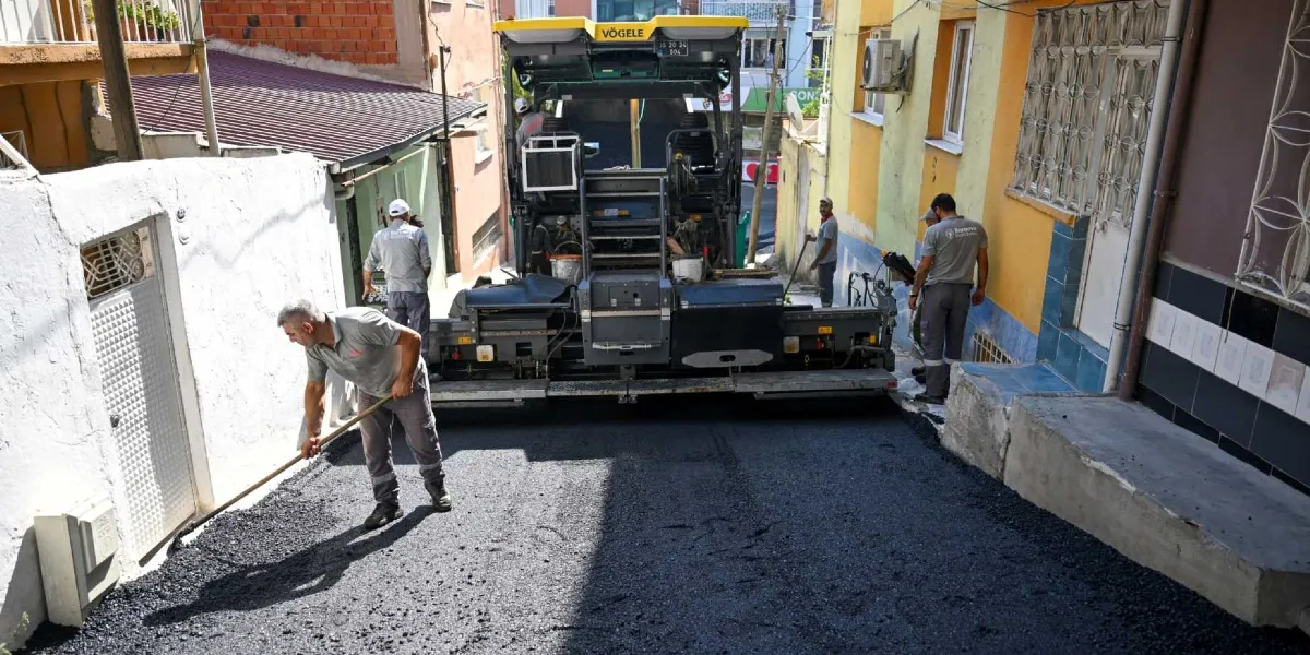 Dar bir sokakta asfalt serme çalışması yapan Bornova Belediyesi ekipleri. Bir işçi elindeki tırmıkla asfaltı düzeltirken, arkasında büyük bir asfalt serme makinesi ve diğer belediye personeli yer alıyor.