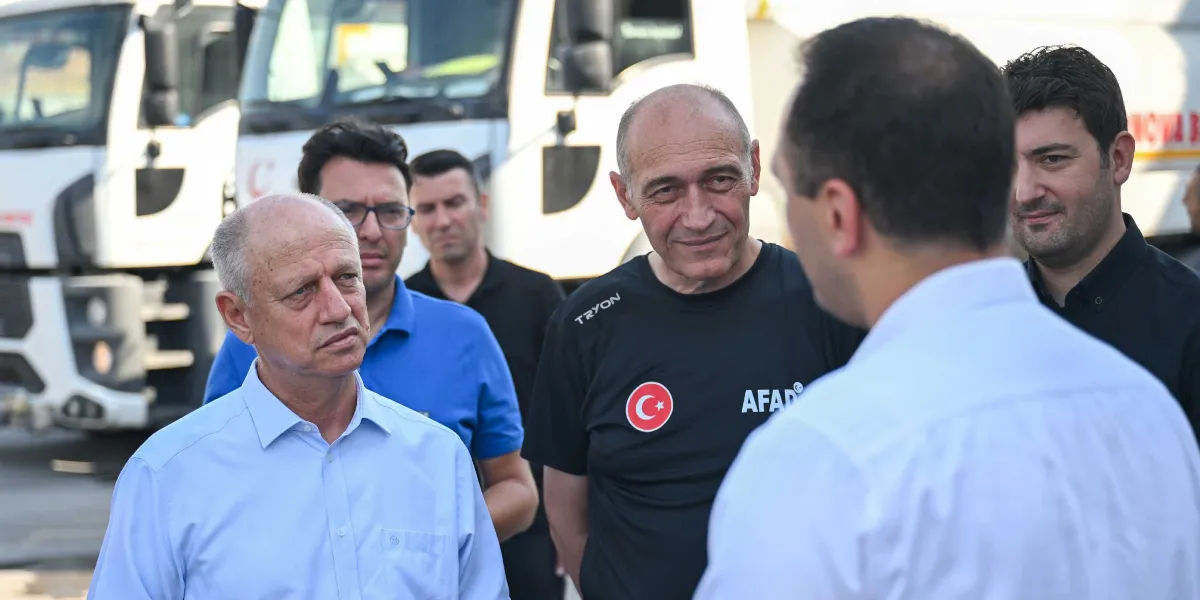 Açık havada, arkalarında beyaz iş makinelerinin bulunduğu bir alanda bir grup erkek bir arada duruyor. Ön planda, üzerinde Türk bayrağı arması ve AFAD yazısı bulunan siyah bir tişört giymiş orta yaşlı bir adam, yanındaki kişilerle konuşuyor. Yanında açık mavi gömlekli bir beyefendi ve diğer yetkililer dikkatle dinliyor.