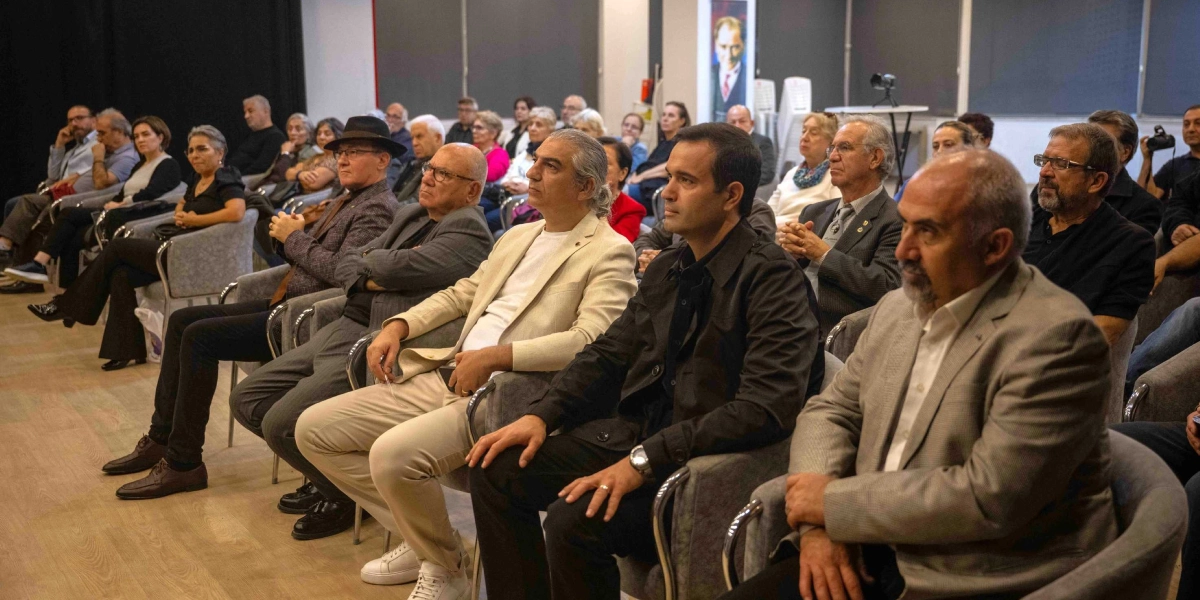 Kapalı bir salonda düzenlenen söyleşide, gri koltuklarda yan yana oturan ve dikkatle dinleyen katılımcılar yer alıyor. Ön sırada, bej ceketli ve beyaz tişörtlü bir adam, yanında koyu renk ceketli bir kişi ve en sağda bej renkli ceket giymiş sakallı bir katılımcı bulunuyor. Arka sıralarda ise farklı yaş gruplarından kadın ve erkeklerden oluşan kalabalık bir izleyici grubu oturuyor. Salonun arka duvarında, Atatürk'ün yer aldığı dikey bir poster asılı duruyor.