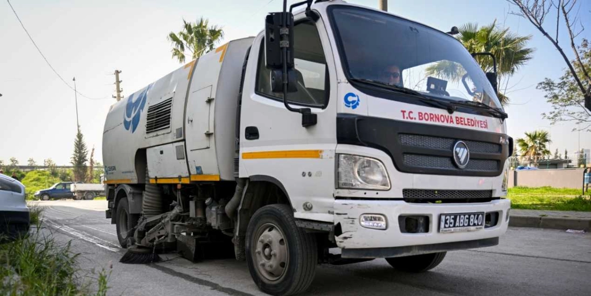 Üzerinde T.C. Bornova Belediyesi yazan, beyaz renkli bir yol süpürme aracı asfalt yolda temizlik çalışması yapıyor.
