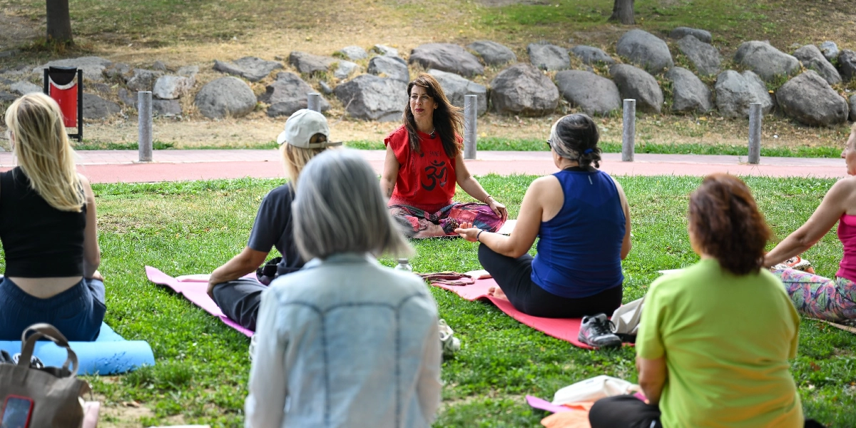 Parkta çimenlerin üzerinde bağdaş kurarak oturan bir grup kadın, yoga veya meditasyon etkinliği için bir eğitmeni dinliyor. Kırmızı tişörtlü eğitmen, grubun önünde oturarak konuşurken, diğer katılımcılar sırtları dönük şekilde matlarının üzerinde onu izliyor. Arka planda taşlarla çevrili bir yürüyüş yolu ve ağaçlık bir alan bulunuyor.