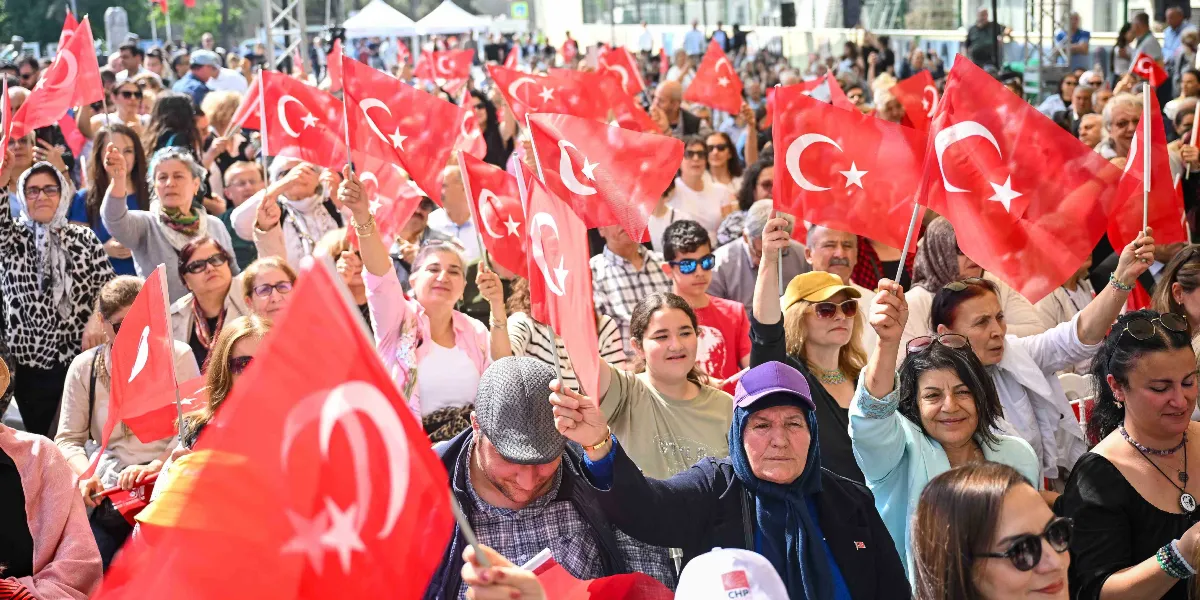 Açık havada düzenlenen bir etkinlikte ellerinde Türk bayrakları tutan kalabalık bir grup insan. Çoğunluğu kadınlardan oluşan katılımcılar, bayrakları havaya kaldırarak coşkulu bir şekilde bir arada bulunuyor. Arka planda ise ağaçlar ve etkinlik alanındaki diğer insanlar seçiliyor.