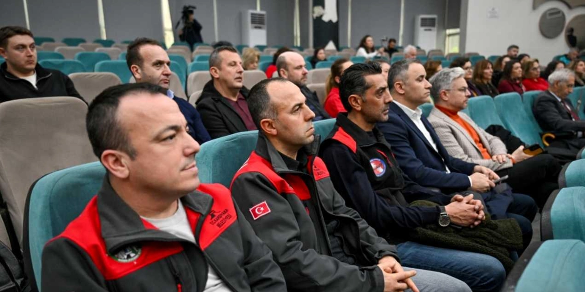 Konferans salonunda yan yana oturmuş, dikkatle dinleyen bir grup insan. Ön planda, üzerinde Karşıyaka Belediyesi arması ve Türk bayrağı yaması bulunan gri-kırmızı iş kıyafetli iki görevli yer alıyor. Arka sıralarda ise takım elbiseli ve günlük kıyafetli katılımcılar bulunuyor. Salonun koltukları turkuaz renginde ve ortamda ciddi bir toplantı havası hakim.