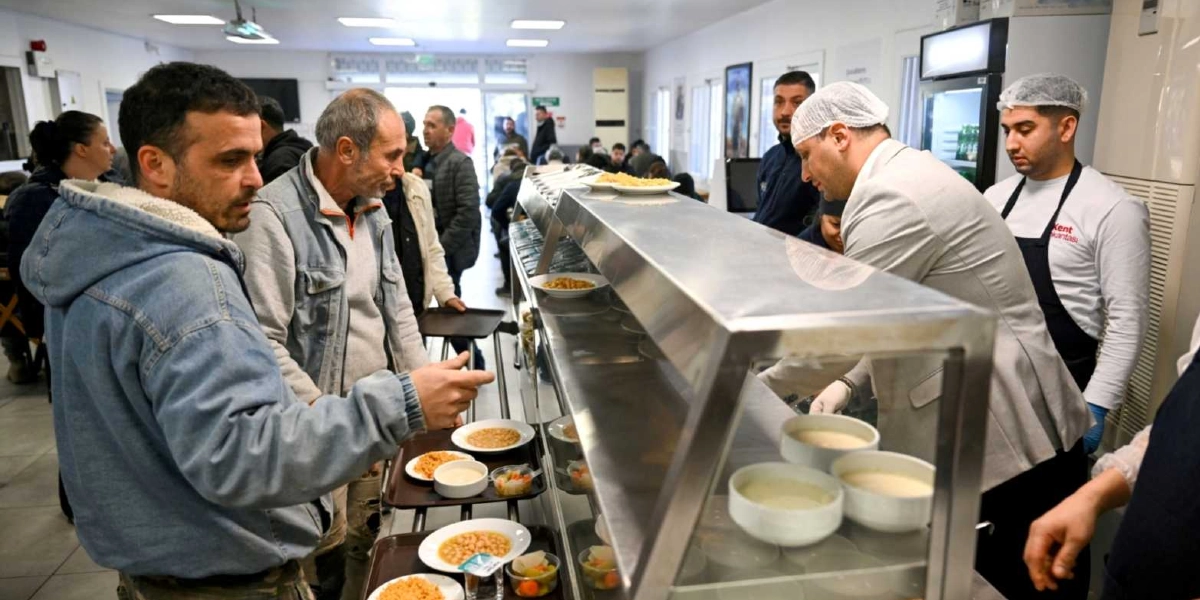 Bornova Belediyesi'ne ait bir aşevinde, yemek dağıtım tezgahının önünde sıra bekleyen vatandaşlar ve servis yapan görevliler. Metal bir tezgahın arkasında, saç bonesi takan görevliler tabaklara yemek koyarken, ön tarafta bekleyen kişiler tepsilerini alarak sırayla ilerliyor. Tezgahın üzerinde çeşitli yemeklerin bulunduğu tabaklar ve kaseler yer alıyor.