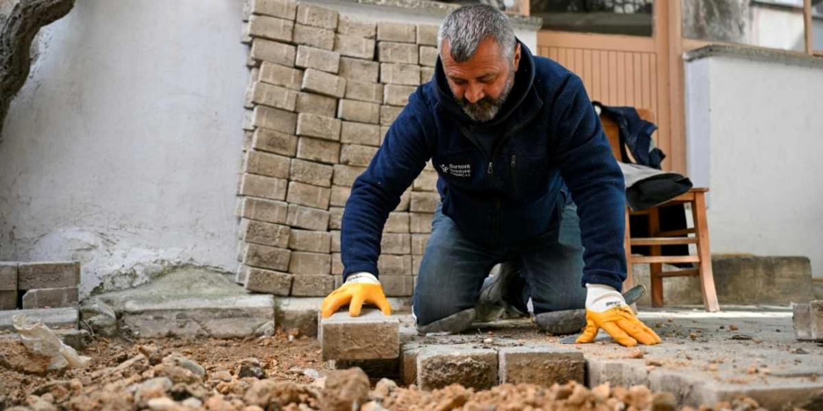 Bornova Belediyesi logolu lacivert bir polar giymiş, sarı iş eldivenleri takan bir belediye çalışanı, dizlerinin üzerine çökmüş vaziyette toprak zemine parke taşları döşüyor. Çalışanın arkasında üst üste dizilmiş parke taşları ve ahşap bir sandalye bulunuyor.