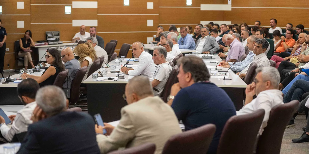 Modern bir toplantı salonunda, uzun masalar etrafında oturmuş, dikkatle dinleyen ve not alan kalabalık bir katılımcı grubu. İnsanlar gündelik kıyafetler içinde, masaların üzerindeki mikrofonlar ve belgeler eşliğinde bir sunumu veya tartışmayı takip ediyor.