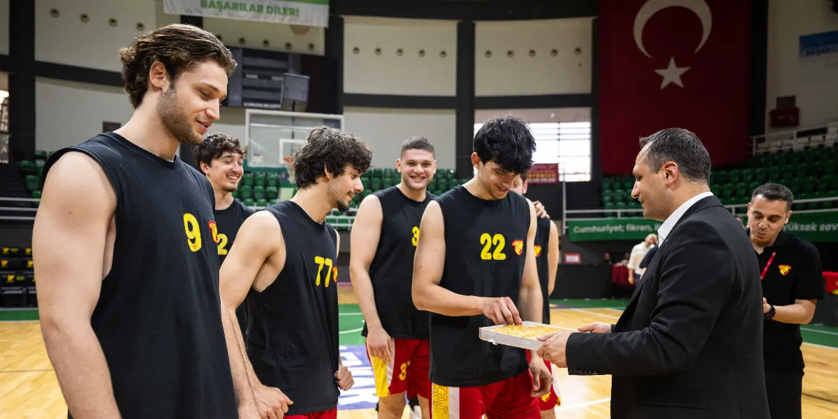 Siyah basketbol formaları giymiş Göztepe oyuncuları, kapalı bir spor salonunda kendilerini ziyaret eden ve elinde bir tepsi baklava tutan takım elbiseli bir yetkiliyle sohbet ediyor. Oyuncular gülümseyerek baklavadan alırken, arka planda asılı büyük bir Türk bayrağı ve tribünler görülüyor.