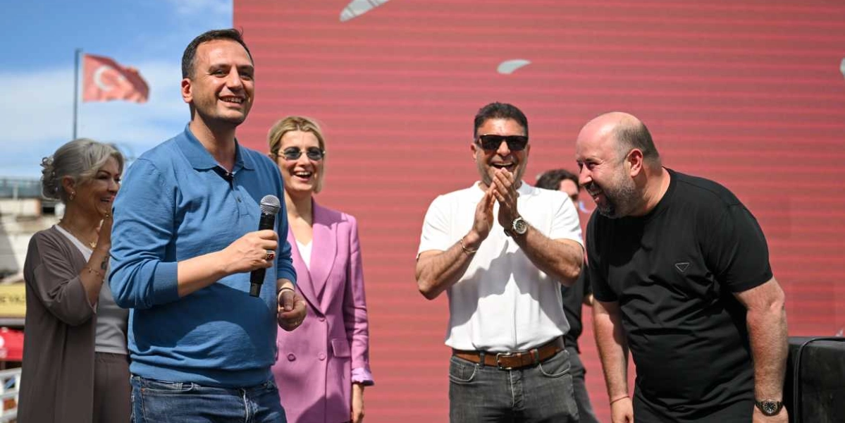 Açık havada, kırmızı bir arka plan önünde duran beş kişilik bir grup, neşeli bir şekilde gülümseyerek bir etkinlikte yer alıyor. En önde, mavi polo yaka tişört giyen bir erkek elinde mikrofonla konuşurken, yanında pembe ceketli bir kadın, beyaz tişörtlü ve güneş gözlüklü bir erkek ile siyah tişörtlü bir erkek ona eşlik ediyor. Arka planda ise dalgalanan bir Türk bayrağı görünüyor.