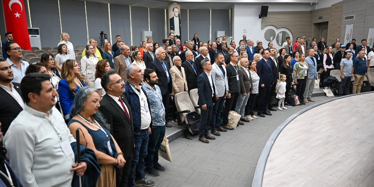 Modern bir konferans salonunda, ayakta duran ve saygı duruşunda bulunan kalabalık bir grup insan. Salonun ön tarafında asılı büyük bir Türk bayrağı ve Mustafa Kemal Atatürk'ün portresi yer alıyor. Katılımcılar arasında farklı yaş ve meslek gruplarından insanlar bulunuyor; bazıları resmi kıyafetler, bazıları ise günlük kıyafetler giymiş durumda. Salonun sahne kısmı boş ve kavisli bir yapıya sahip.