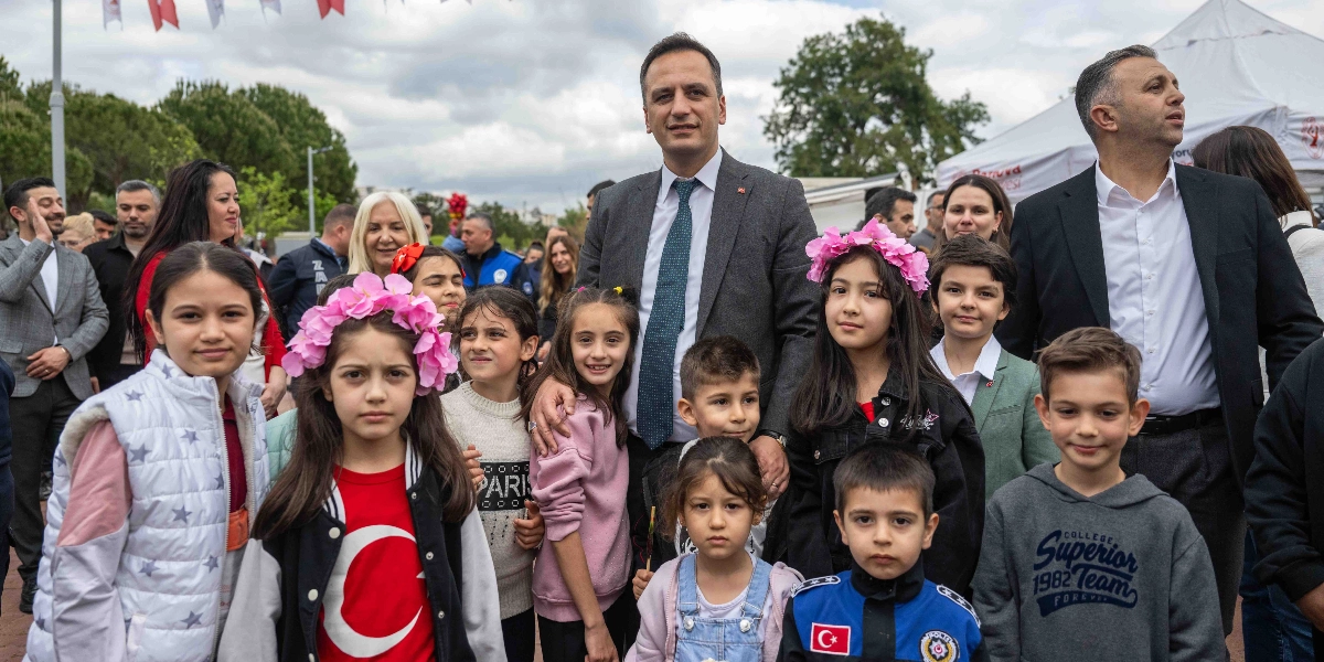 Açık havada düzenlenen bir etkinlikte, takım elbiseli bir erkek ve bir grup çocuk yan yana durarak objektife gülümsüyor. Çocuklardan bazıları başlarında pembe çiçekli taçlar takarken, bir çocuk üzerinde Türk bayrağı baskılı tişört, bir diğeri ise üzerinde Türk bayrağı arması bulunan polis üniforması giyiyor. Arka planda ise etkinliğe katılan diğer yetişkinler ve beyaz bir çadır yer alıyor.