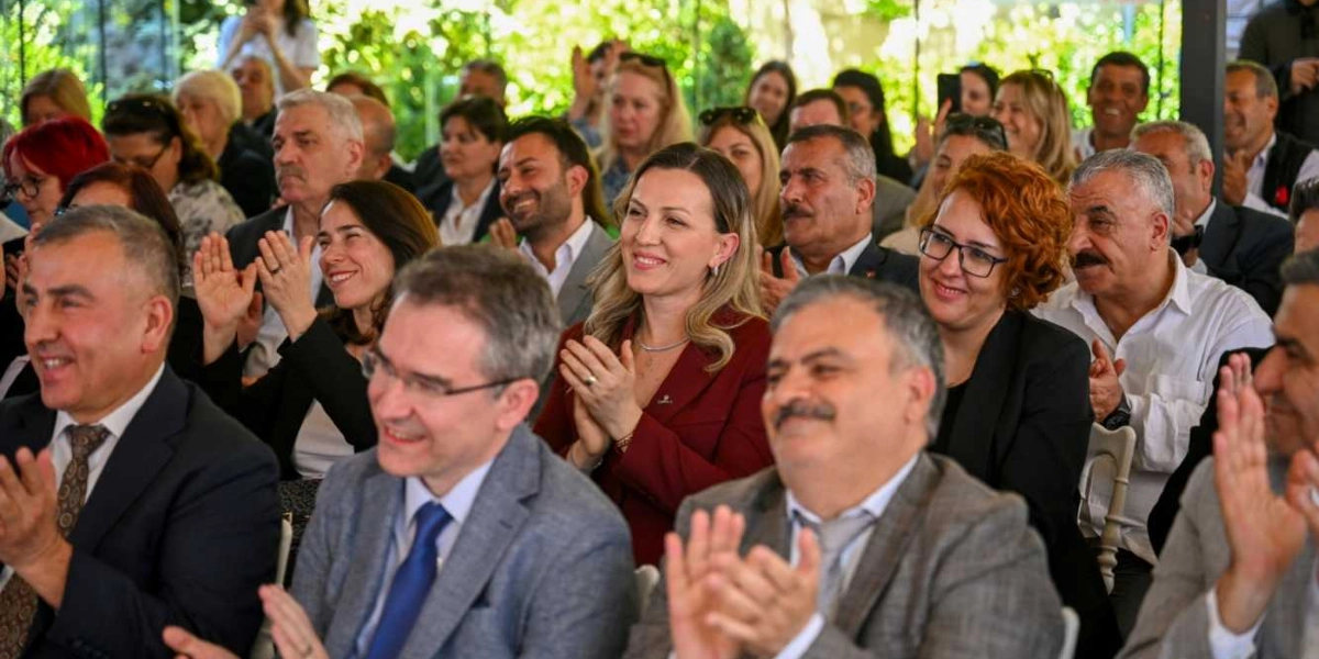 Açık havada düzenlenen bir etkinlikte, katılımcıların bir konuşmayı veya performansı alkışladığı an. Ön planda takım elbiseli erkekler ve bordo ceketli bir kadının yer aldığı, çeşitli yaş gruplarından oluşan kalabalık bir grup, gülümseyerek ve elleriyle alkış tutarak etkinliği takip ediyor. Arka planda ise ağaçların gölgesinde oturan diğer katılımcılar görülüyor.