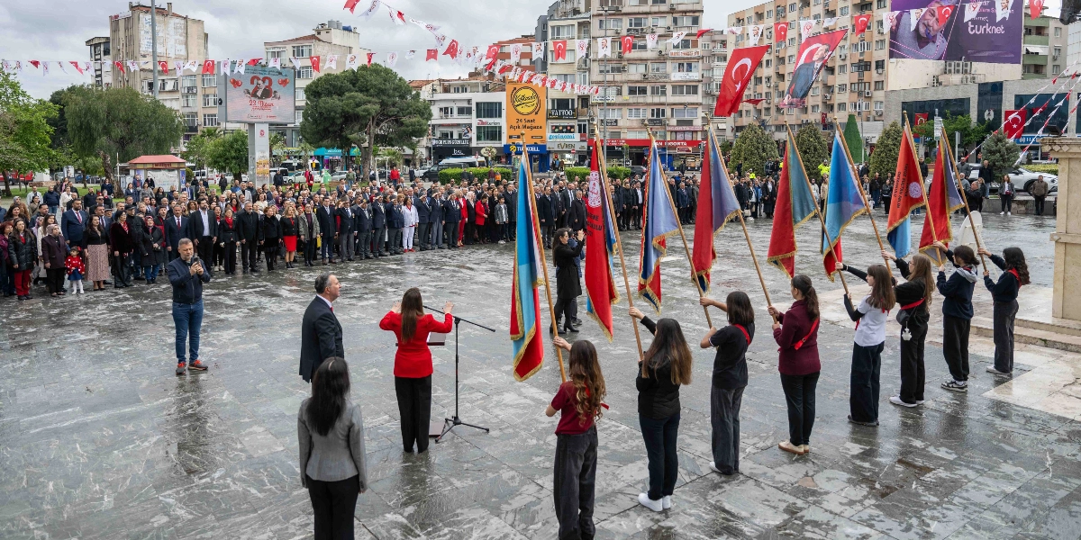 Bornova'da bir meydanda düzenlenen 23 Nisan Ulusal Egemenlik ve Çocuk Bayramı töreninde, ellerinde okul flamaları ve Türk bayrakları taşıyan bir grup öğrenci tören alanında sıralanmış durumda. Meydanın arka kısmında resmi kıyafetli protokol üyeleri ve vatandaşlar töreni izliyor. Ön planda, bir kürsüde konuşma yapan kırmızı ceketli bir kadın ve yanında duran bir erkek yetkili bulunuyor. Çevredeki binalar Türk bayraklarıyla süslenmiş ve gökyüzü bulutlu görünüyor.