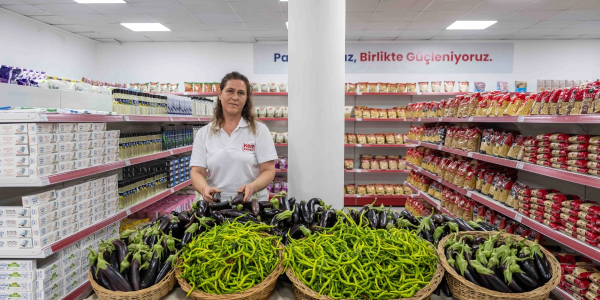 Beyaz polo yaka üniformalı bir kadın, market reyonlarının önünde durarak elindeki patlıcanları düzenliyor. Önündeki tezgâhta hasır sepetler içinde taze patlıcanlar ve sivri biberler bulunuyor. Marketin arka duvarında 