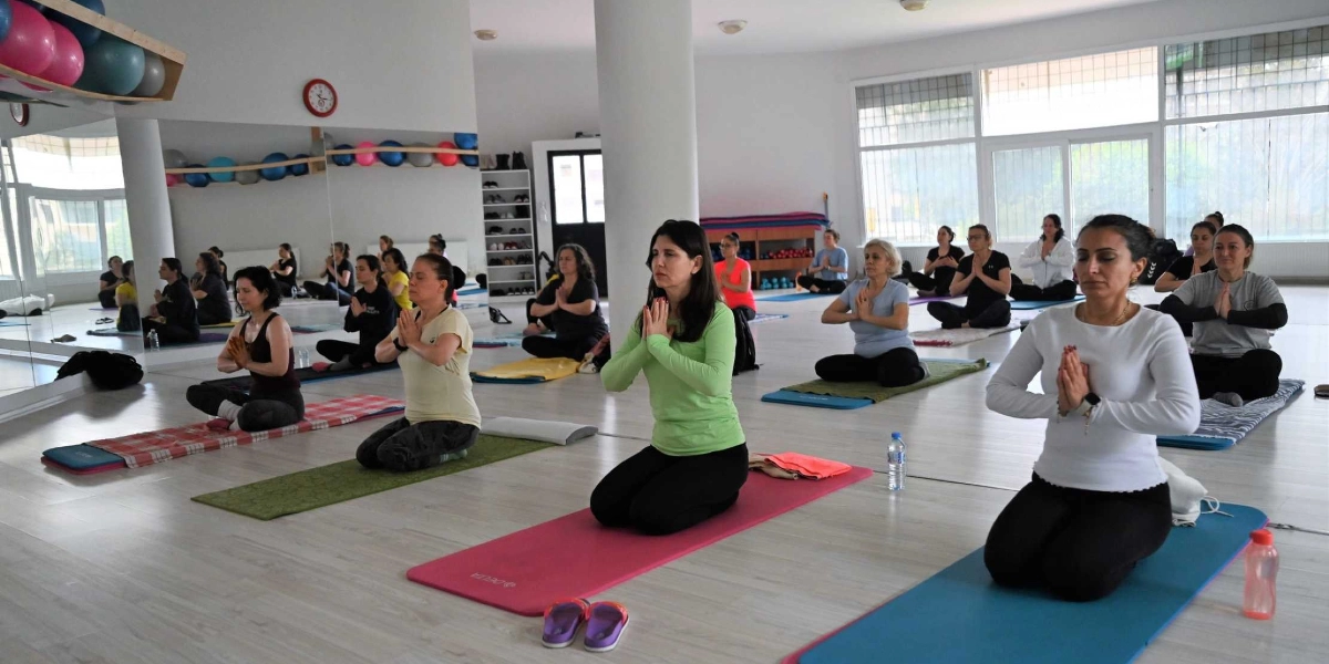 Aydınlık ve geniş bir spor salonunda, yoga matları üzerinde diz çökmüş şekilde oturan ve ellerini göğüs hizasında birleştirerek meditasyon yapan kadınlar grubu. Katılımcılar rahat kıyafetler giymiş olup, salonun duvarında bulunan büyük aynalar sayesinde grup üyelerinin yansımaları da görülmektedir.