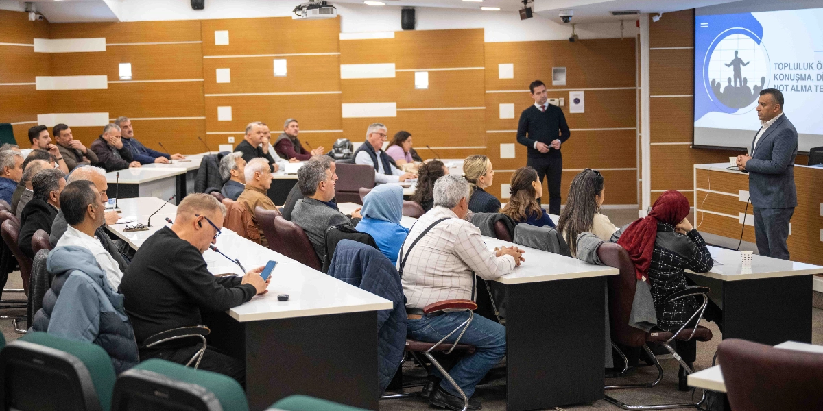 Bornova Muhtarlar Akademisi kapsamında düzenlenen bir eğitim toplantısında, takım elbiseli bir konuşmacı katılımcılara hitap ediyor. Modern bir konferans salonunda, sıralarda oturan çok sayıda muhtar dikkatle sunumu dinliyor. Salonun ön tarafındaki büyük ekranda 